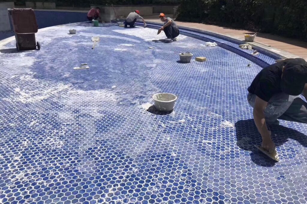 How to Choose Pool Mosaic Tiles: A Project-Based Guide 8 Installers setting and grouting blue hexagonal mosaics across a swimming pool floor