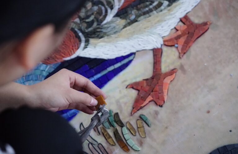 Craftsperson hand-cutting mosaic pieces beside a bird motif at JNJMosaic