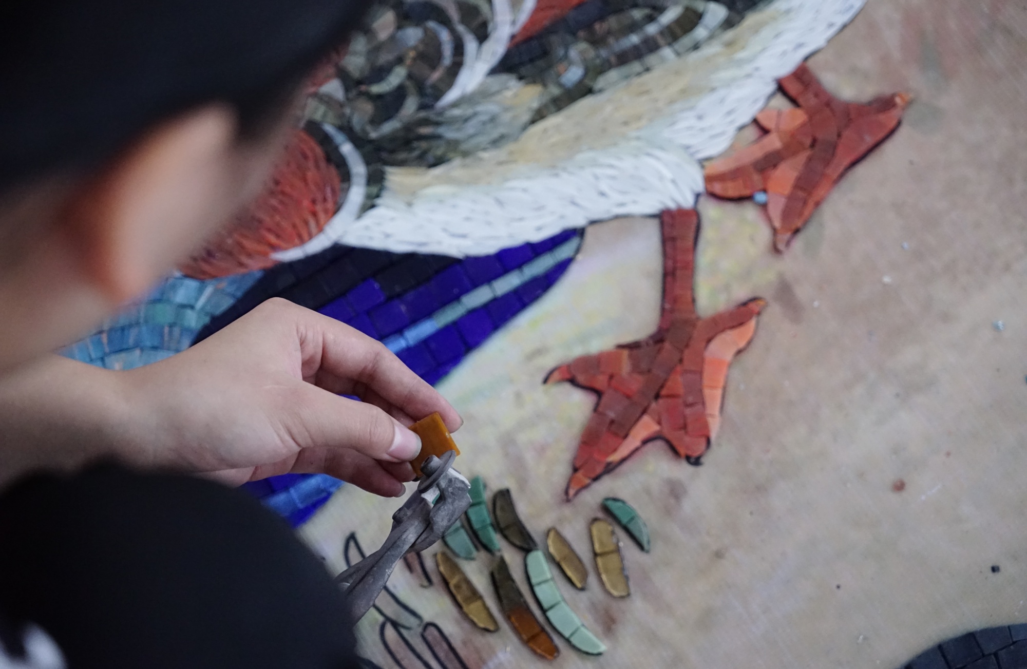 Craftsperson hand-cutting mosaic pieces beside a bird motif at JNJMosaic